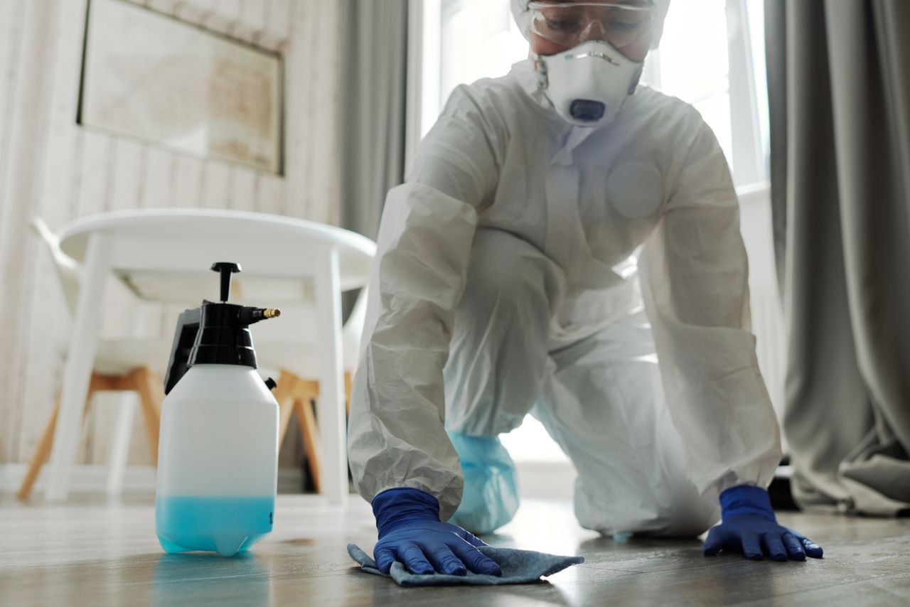 mobile-02 A person wearing protective equipment is cleaning an indoor floor with disinfectant spray and a cloth.